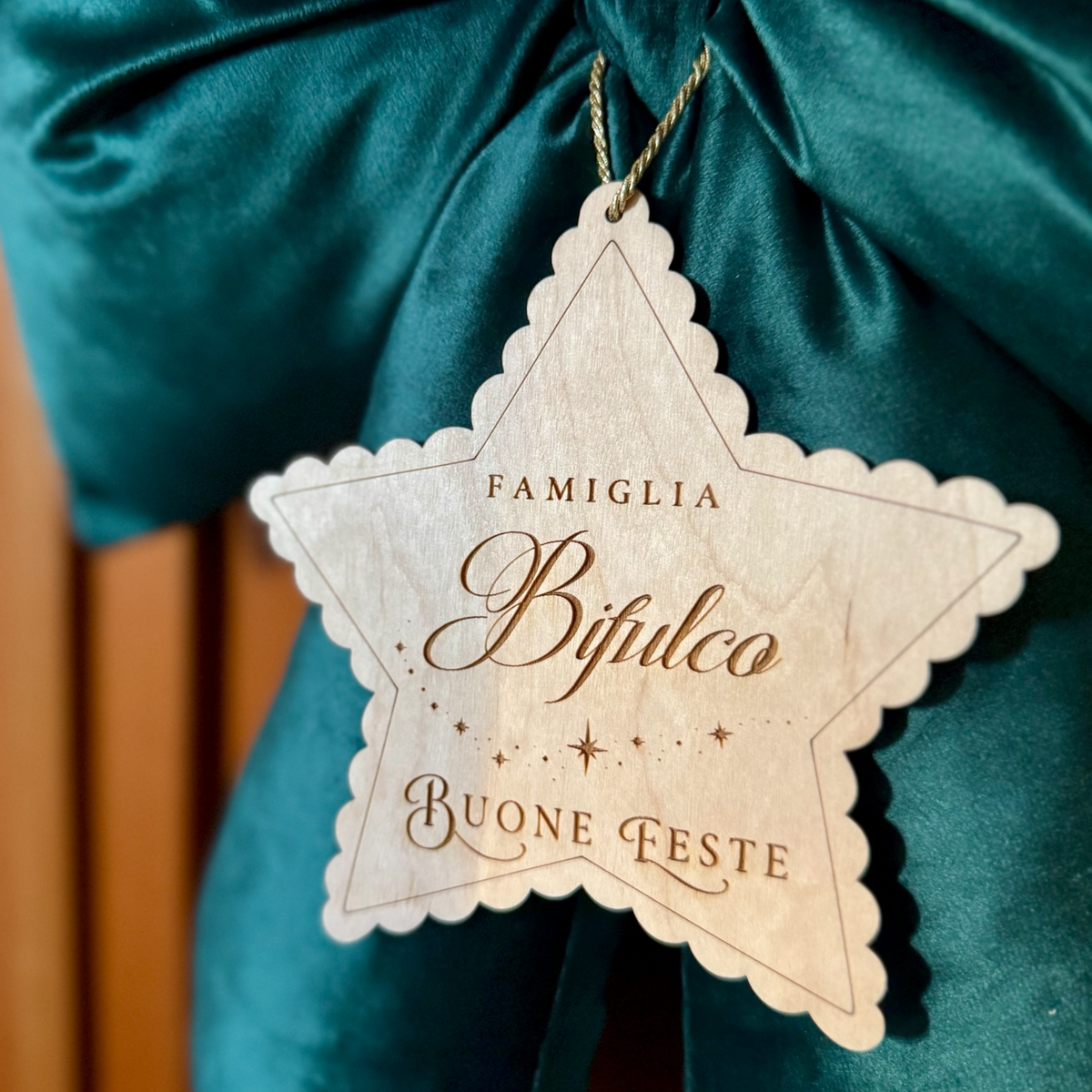 Dettaglio della targhetta a forma di stella in legno con cognome della famiglia inciso e scritta Buone Feste, sul fiocco natalizio verde.